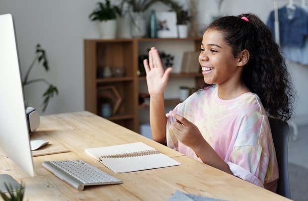 Girl sitting at desk in bedroom waving at computer.