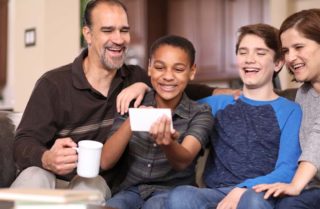 Mother, father, and two boys sitting on couch looking at cell phone, all smiling.