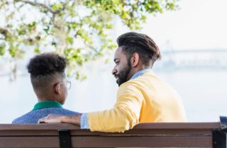 An adult and child sit on a bench, with the child looking out at a lake and the adult smiling at the child