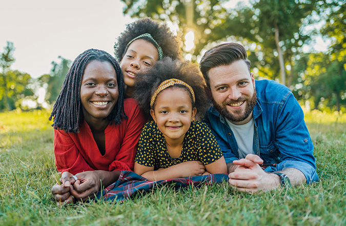 A family of four smiles at the camera while posing for a picture laying on the grass in their backyard. 