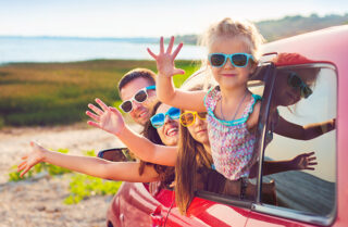 Family of four wearing sunglasses and waving out of a car window on a sunny day