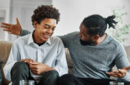 One Black male teenager and one Black male adult are smiling and talking on a couch.