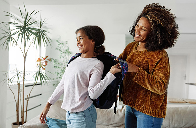 A parent helps her child put on her backpack to go to school.