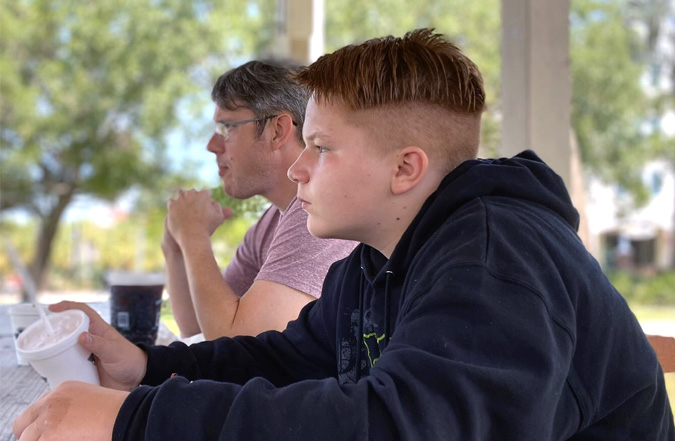 A father, Brian, and his son, 13-year-old Logan, are sitting at a picnic table at a state park, staring out at the scenery.