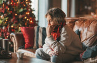 A woman sits on her couch in a living room decorated for the winter holidays, with a contemplative expression.