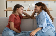 A mother and teenage girl are sitting on the couch, smiling, and engaged in a conversation.