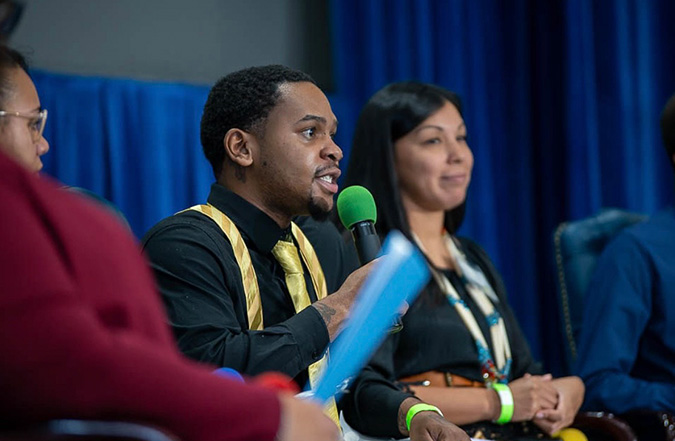 Jamarri Kane-White, a young Black man, sits at a table with fellow panel guests and is holding a microphone as he addresses an audience question.