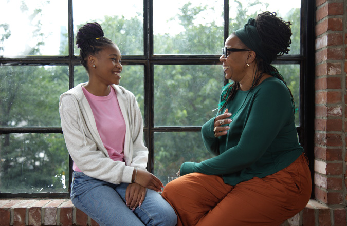 Mother and daughter sitting together and smiling in front of a window.