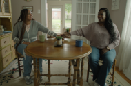 A young woman and her mom sit across from each other at a dining table and hold hands.