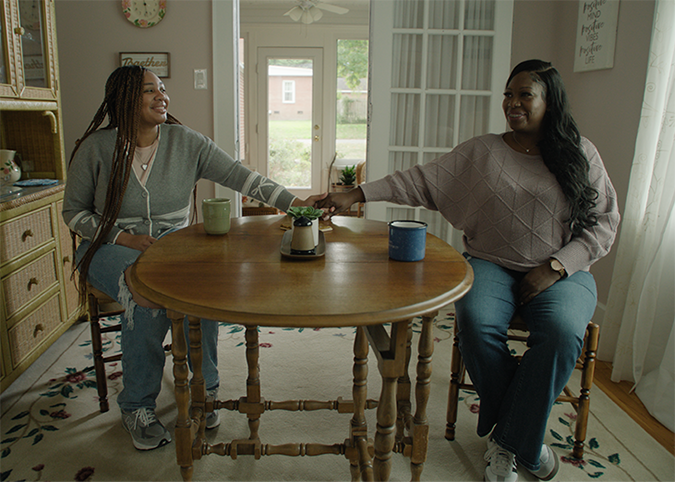 A young woman and her mom sit across from each other at a dining table and hold hands.