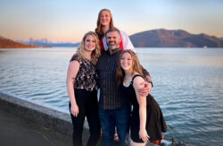 The Hagelman family poses for a photo in front of a body of water and mountains.