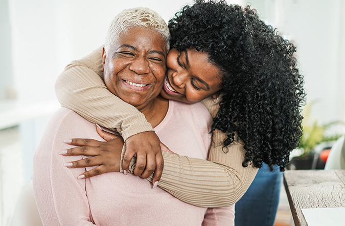 A young woman hugs an older adult who smiles expresses warmth and joy.